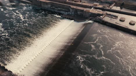 aerial-drone-shot-of-a-river-flowing-over-a-lock-and-dam-in-a-downtown-city-during-golden-hour