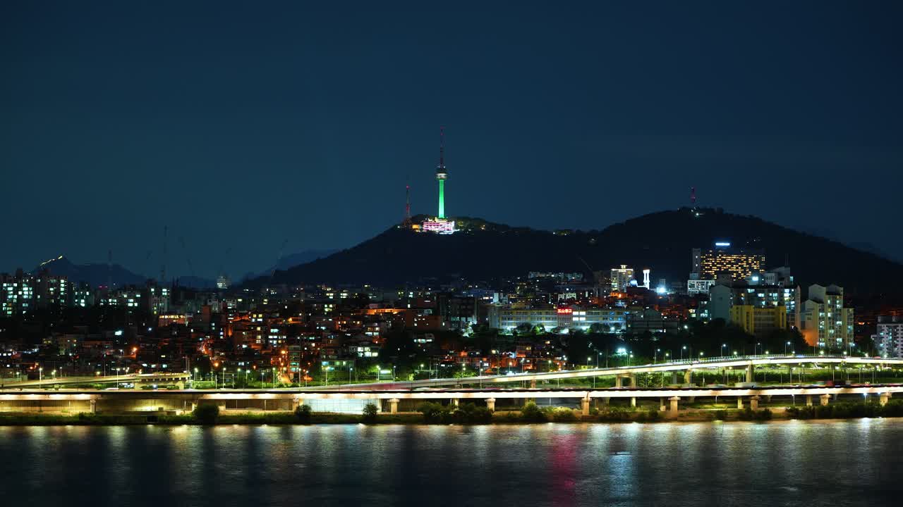 A static time-lapse captures the iconic N Seoul Tower glowing green atop Namsan mountain, overlooking the illuminated Yongsan district and light trails of fast-moving traffic along the Han River