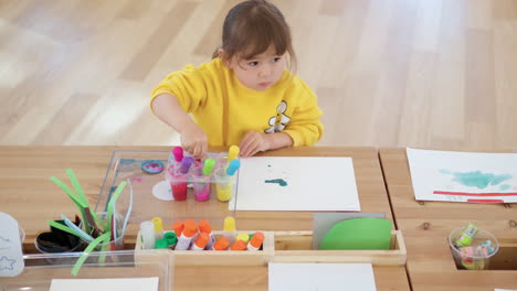 Blond-3-Year-Old-Girl-Study-Dropper-Painting-Sitting-By-the-Desk,-Sucks-in-Colored-Water-into-Pipette-From-Containers