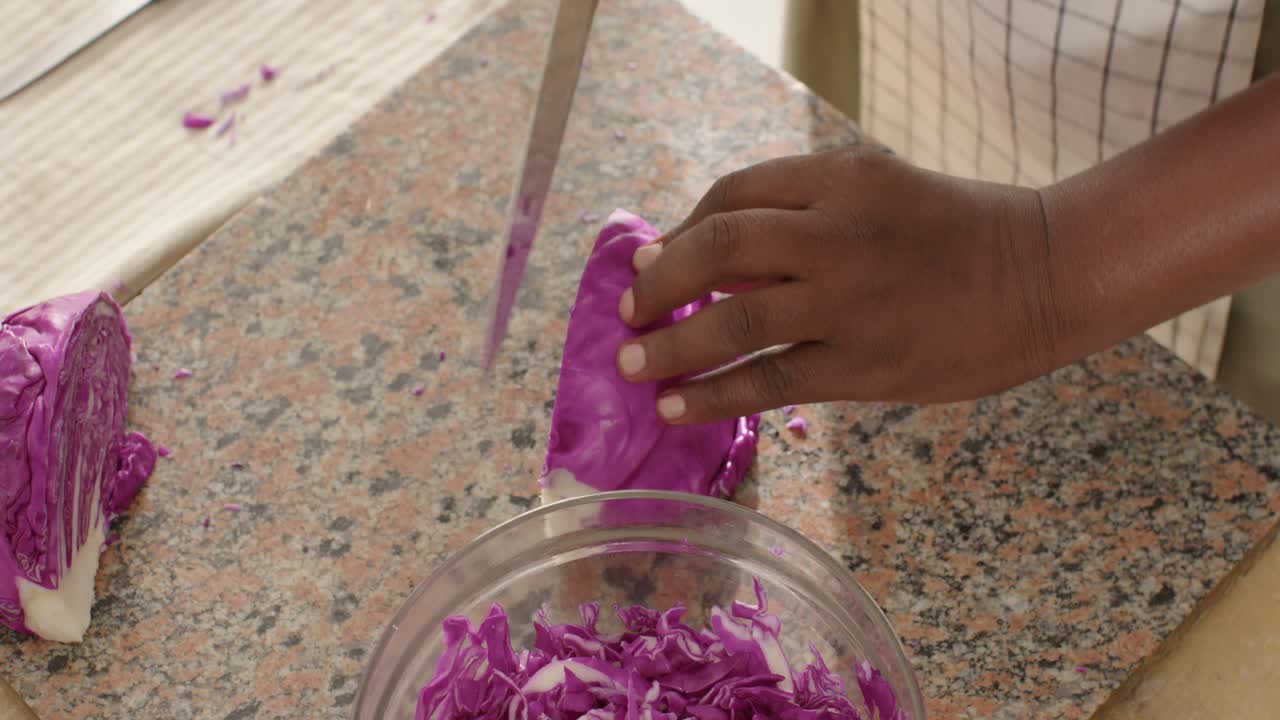 African man in apron placing halved purple cabbage on counter, shredding with chef knife for salad