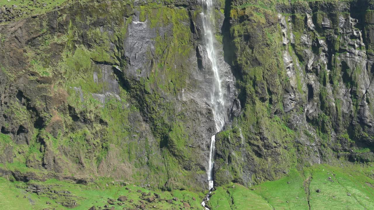 teleobjetivo acercándose a las cascadas de drifandi desde el otro lado de una pradera de hierba verde abierta en islandia