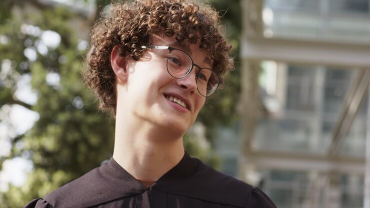 When presenter extends ribboned diploma, male graduate reaching, holding cap and smiling on campus