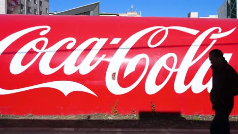 On a bright day, people walk past a wall in Ulaanbaatar, Mongolia, displaying the Coca-Cola logo and its signature red color, advertising the renowned American soft drink.