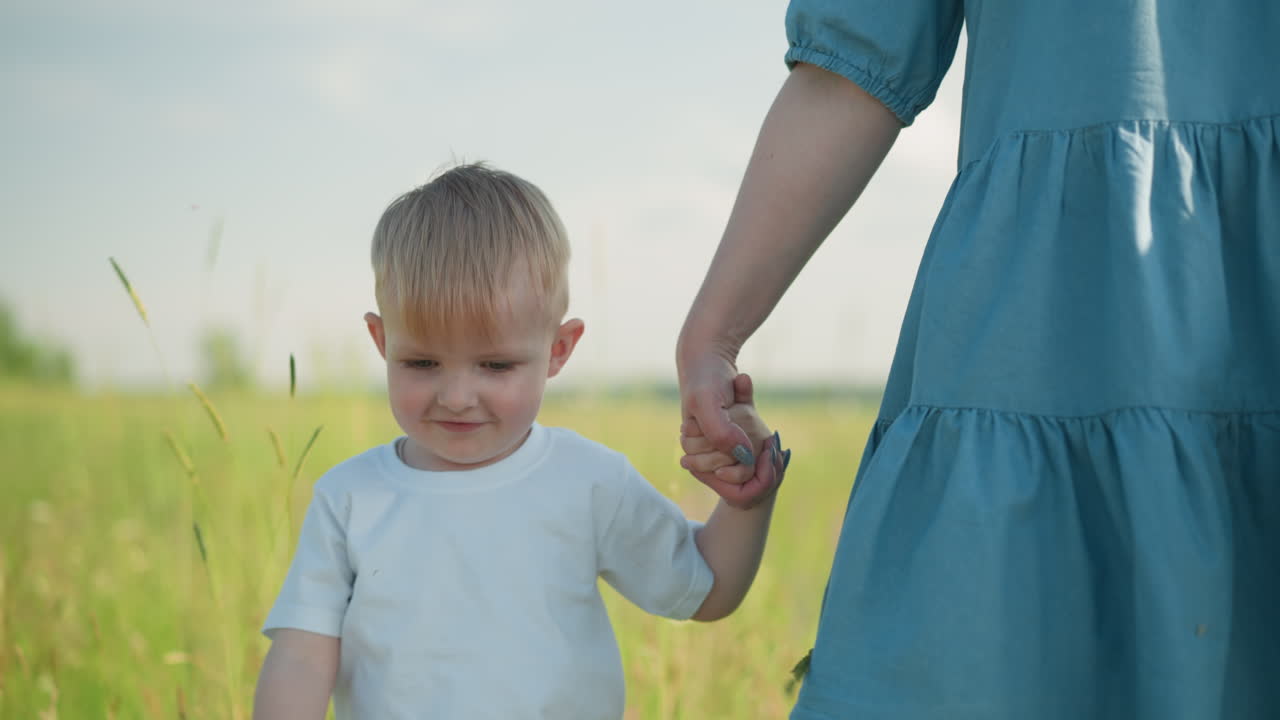 A close-up of a young boy in a white shirt smiling as he walks through a grassy field, holding his mother's hand. The mother, dressed in a blue gown, has her face out of view