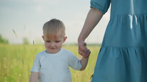 un primer plano de un niño en una camisa blanca sonriendo mientras camina a través de un campo cubierto de hierba, sosteniendo la mano de su madre. la madre, vestida con un vestido azul, tiene su cara fuera de la vista