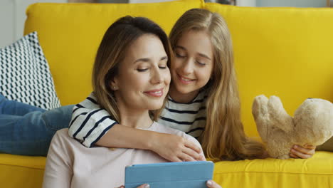 blonde mother and daughter resting on the sofa at home and watching something on a tablet