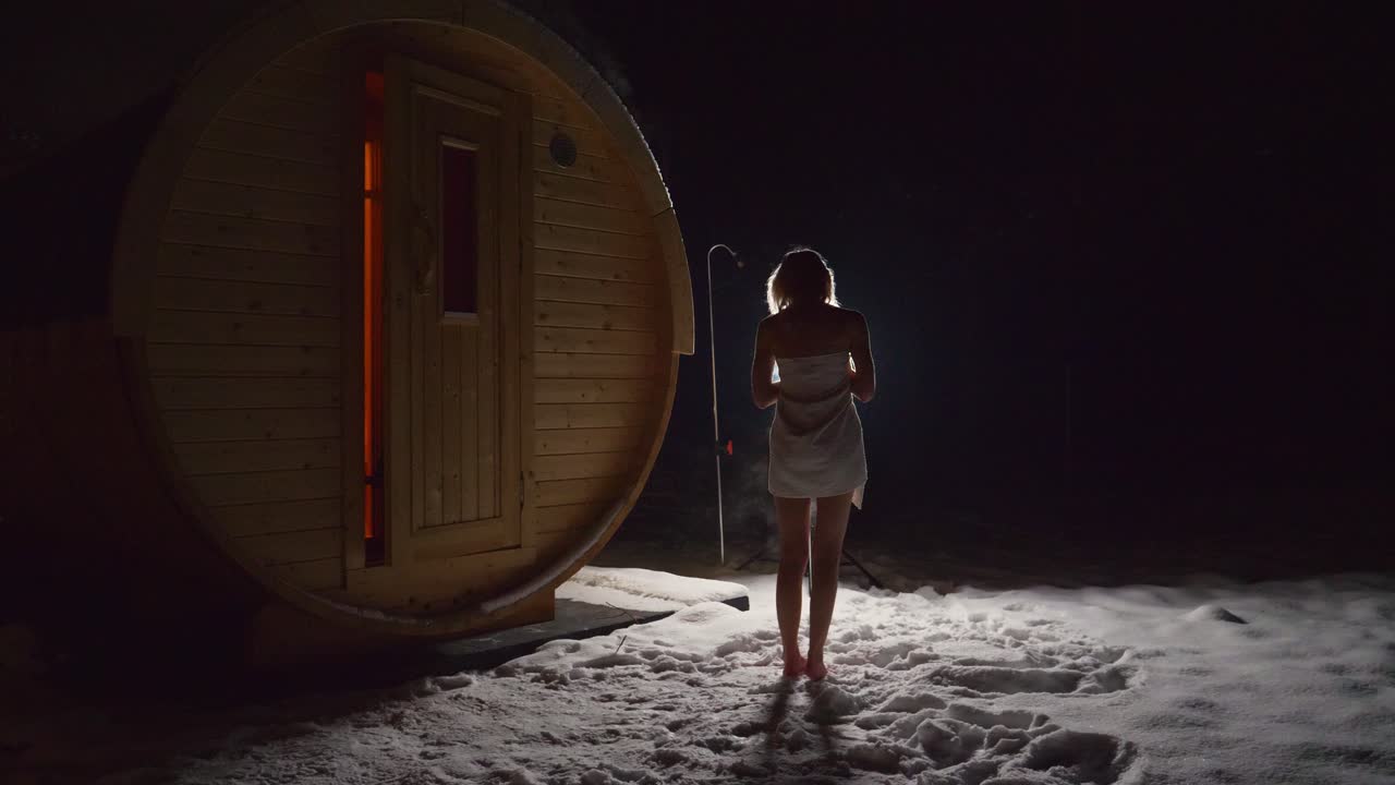 una joven delgada con una toalla parada en la nieve afuera de una casa de sauna