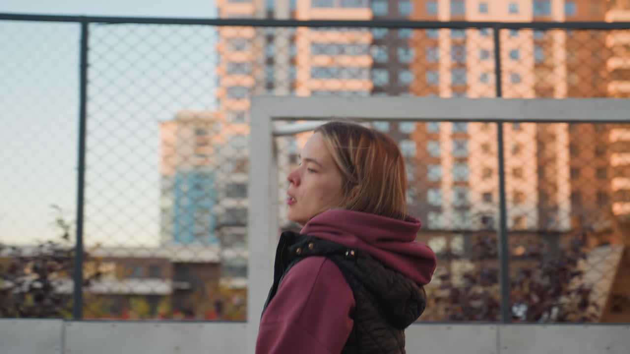Trainer jogging sideways round outdoor sports court in urban environment surrounded by modern high rise apartments under clear sky engaging dynamic lateral movement while wearing hoodie and vest