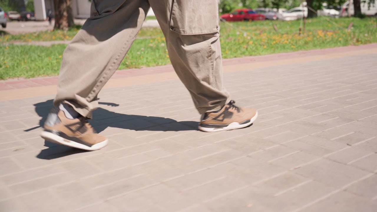 Close-up kaki pria berjalan di tanah beraspal dengan sinar matahari membentuk bayangan. Pria itu mengenakan celana cokelat dan sepatu kets cokelat muda bergerak mantap melalui lingkungan luar ruangan yang menampilkan rumput hijau.