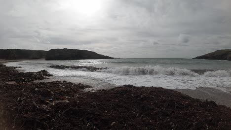 Shimmering-ocean-waves-washing-onto-seaweed-covered-beach-sand-with-islands-on-horizon-Slow-motion