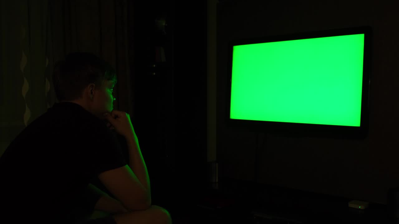 Side view of a young man watching TV with chromakey while sitting on a couch in the living room. Concept. Male watching TV with green screen late at night