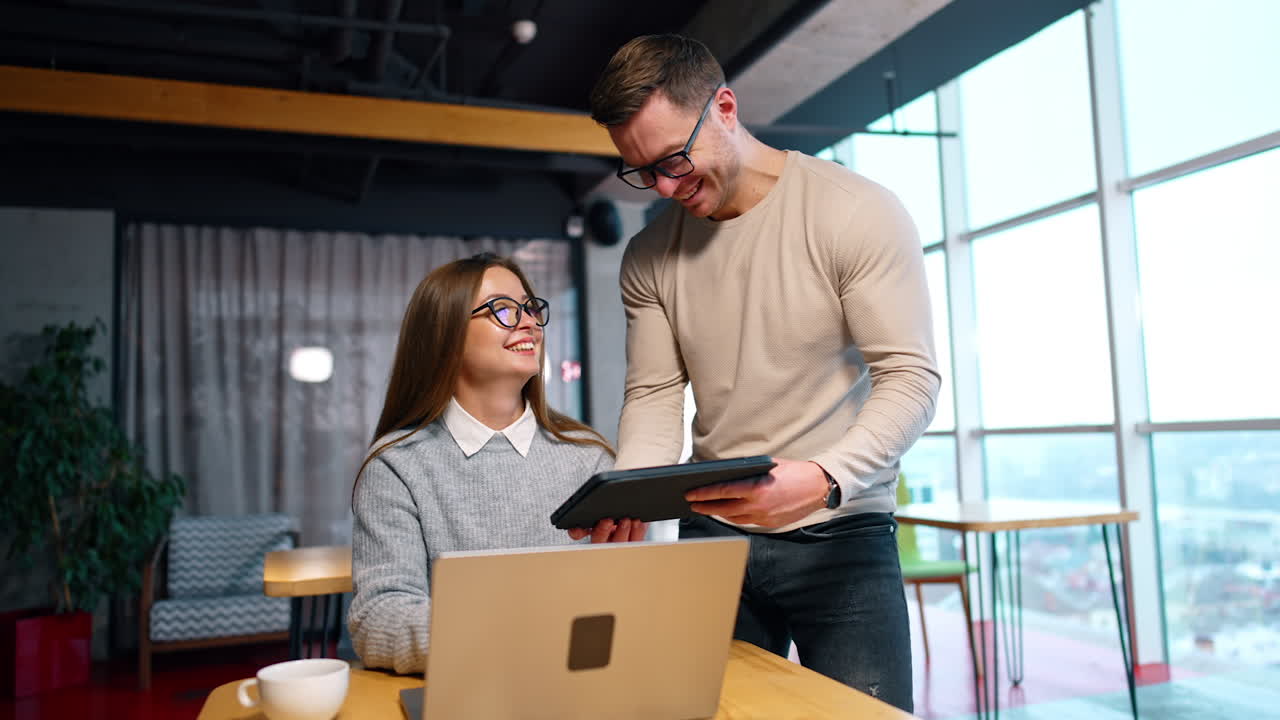 Smiling man in glasses stands near the female colleague. Male employee showing his I-pad to a woman sitting at laptop.