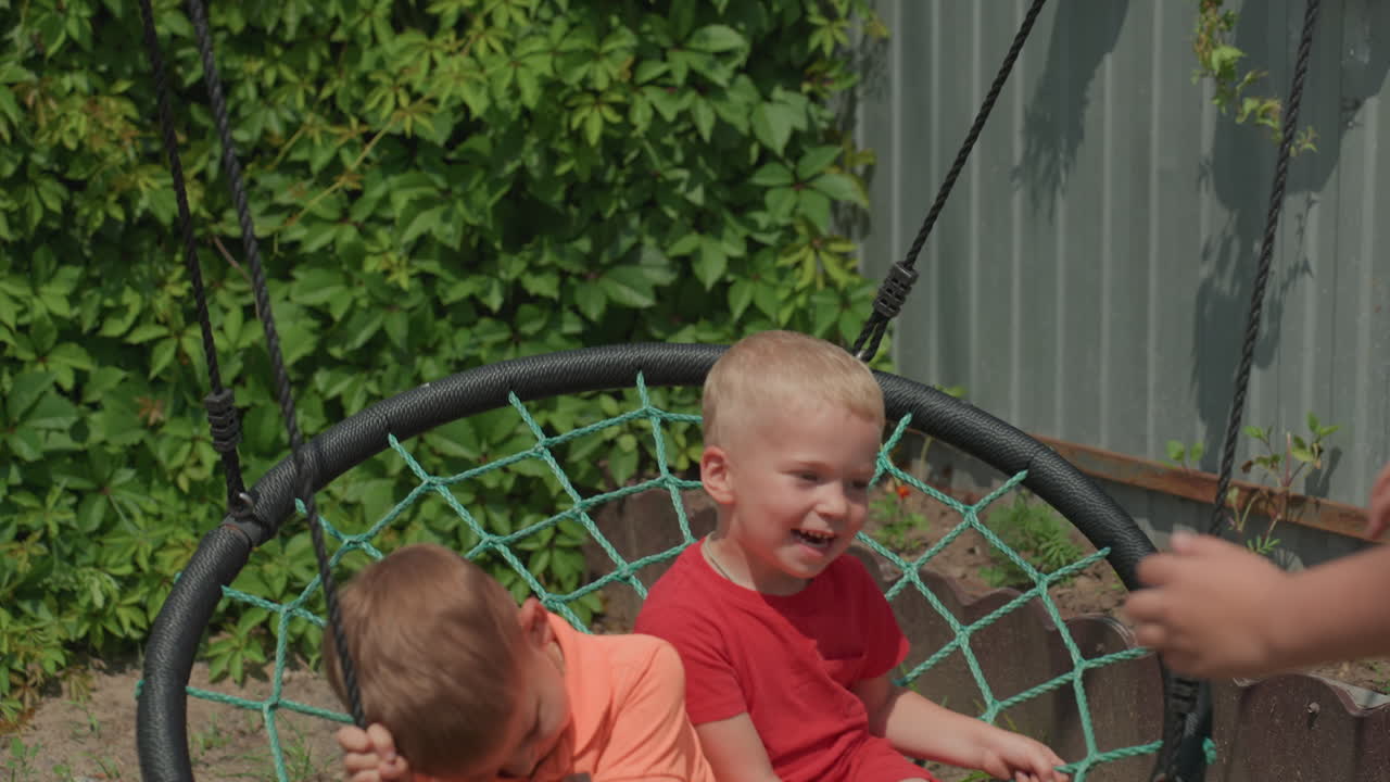 Young Children Laugh On Colorful Swing Outside, Toddlers Enjoy Lively Play On Round Swing With Caregiver Nearby, Little Ones Giggle And Sway On Circular Swing As Adult Supervises In Sunny Backyard