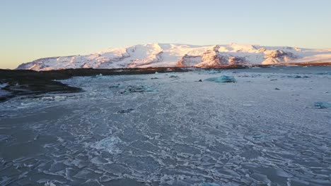 Aerial-Dolly-Drone-Shot-Across-Frozen-Lake-of-Ice-with-Mountainous-Background-Covered-in-Snow,-Iceland