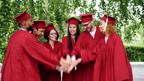 happy graduating students multiethnic group is putting palms together then clapping hands celebrating successful graduation, people are wearing gowns and mortar-boards.