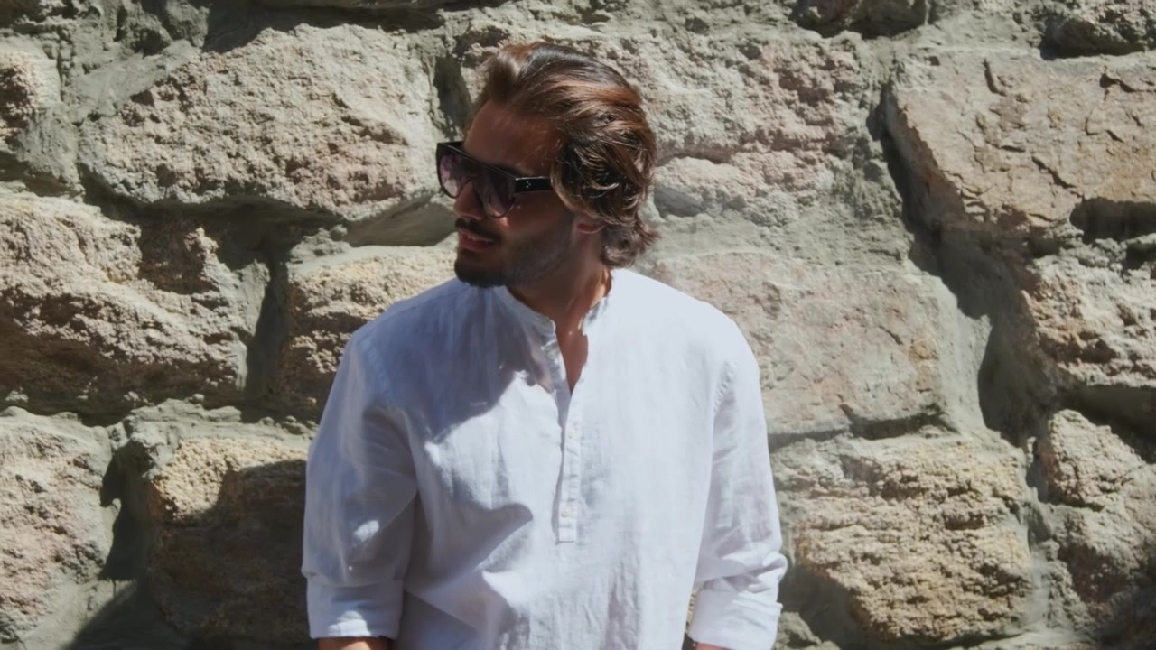 Giovane uomo sicuro di sé che si passa una mano tra i capelli contro un muro di pietra.