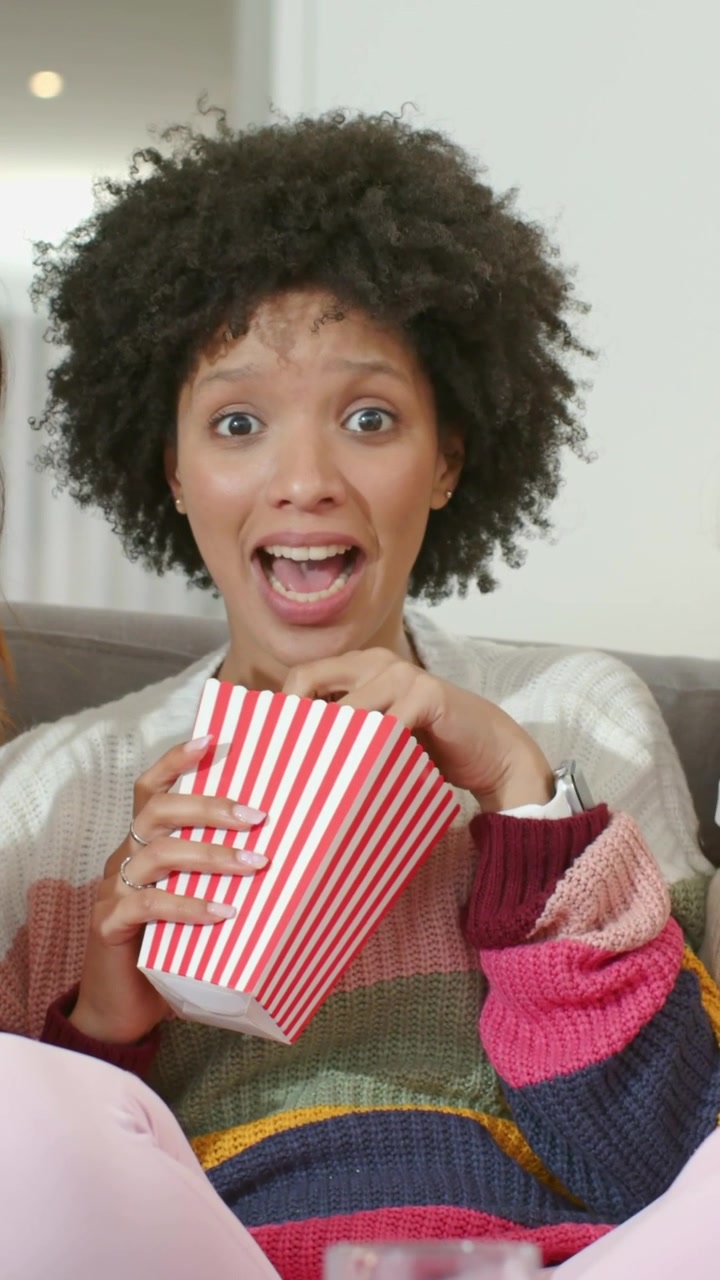 Video verticale: Donna con maglietta a righe sul divano, davanti alla TV, passa dallo shock al sorriso mentre mangia popcorn.