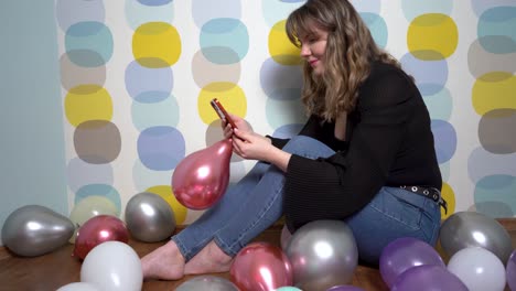 mujer joven sentada alrededor de globos y sosteniendo un teléfono en la mano contra un fondo colorido