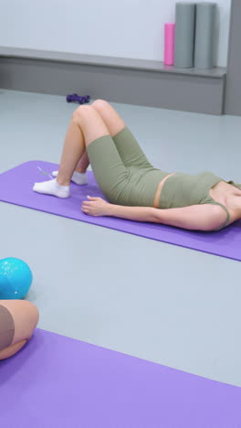 mujer acostada en una alfombra púrpura ajustando su cabello mientras su amiga se arrodilla cerca, observando, globos de colores llenan el fondo, añadiendo un toque lúdico a esta escena de fitness o relajación