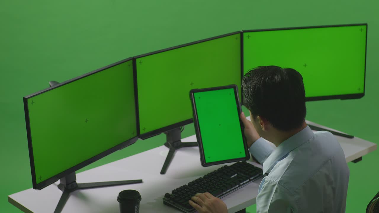 vista lateral de un hombre asiático trabajando con una tableta y varios monitores de computadora con una maqueta de pantalla verde en un estudio de pantalla verde