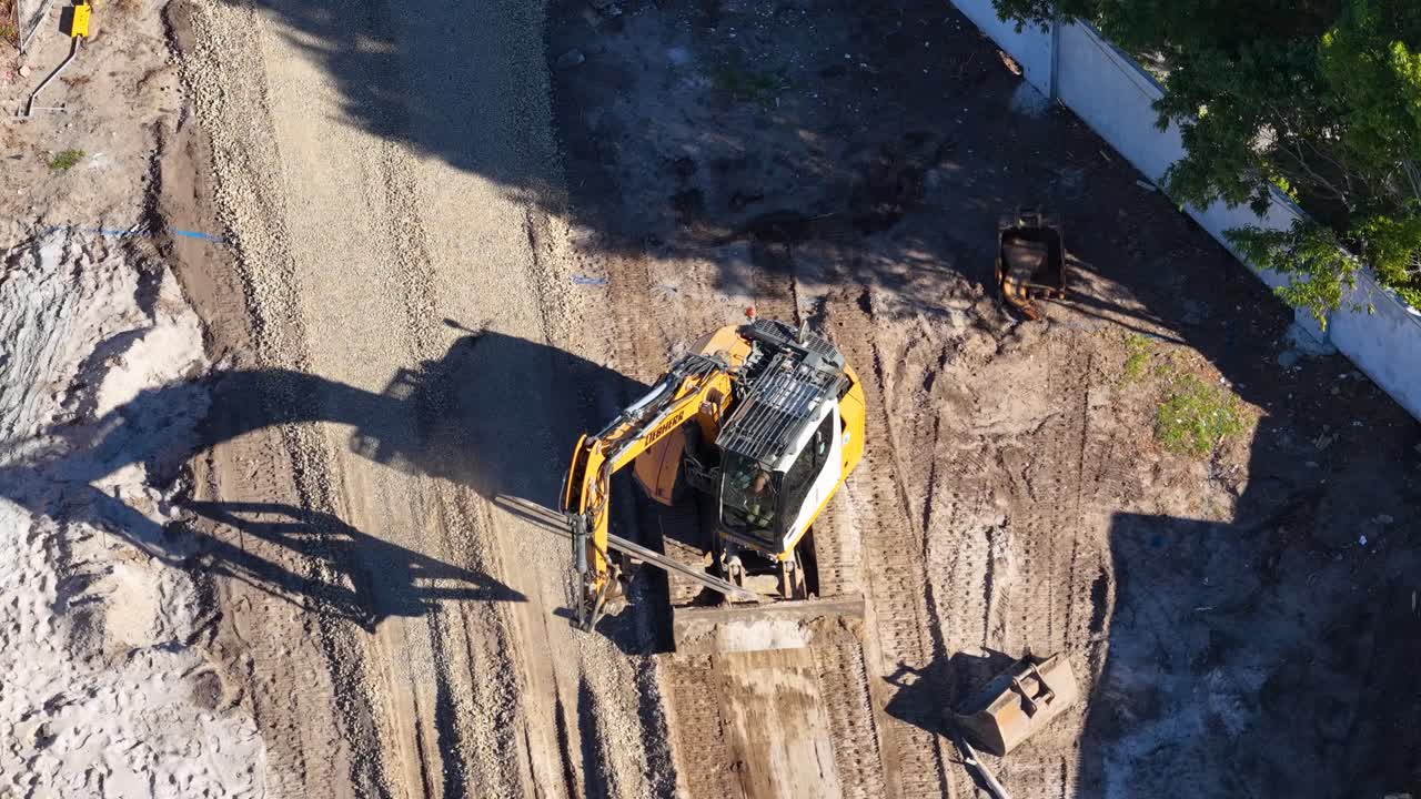 Excavator shifts soil on construction site, overhead drone shot, strong daylight, clear shadows