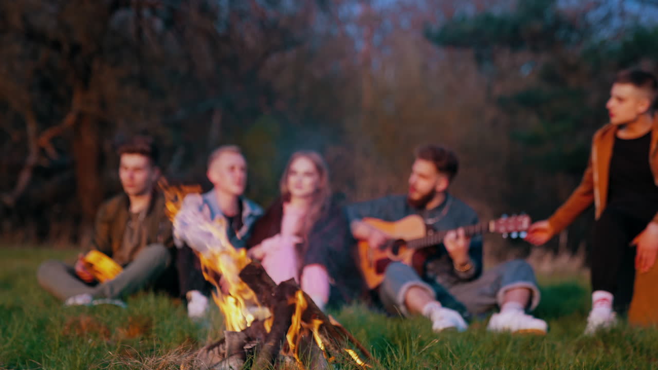Group of friends near the campfire. Young people spending happy time together while sitting near the bonfire in the evening. Young man playing the guitar in nature.