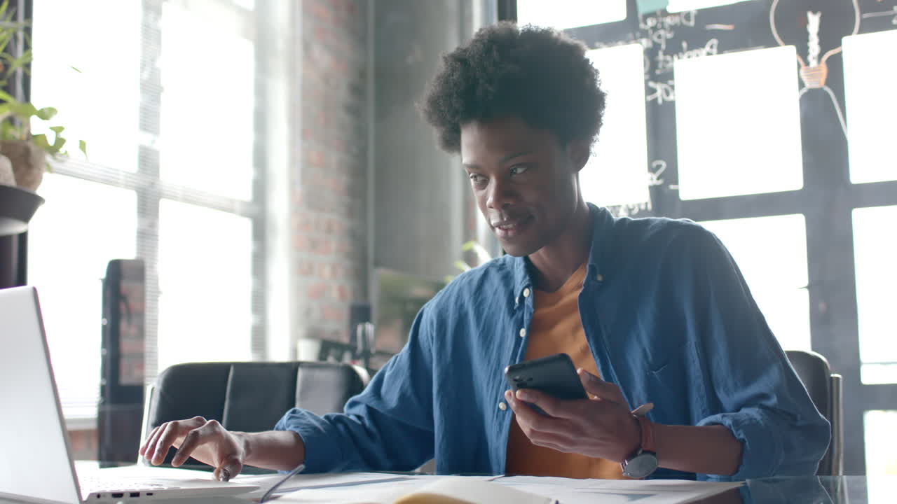 African american casual businessman sitting at desk using smartphone and laptop at home, slow motion