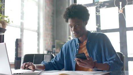 african american casual businessman sitting at desk using smartphone and laptop at home, slow motion