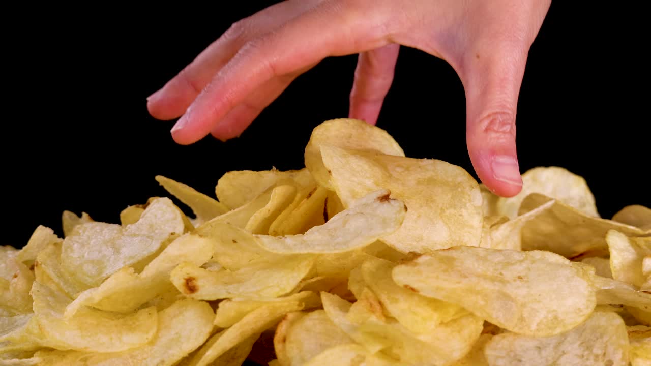 Human hand reaches into pile of potato chips, brightly lit against clean black backdrop, close-up
