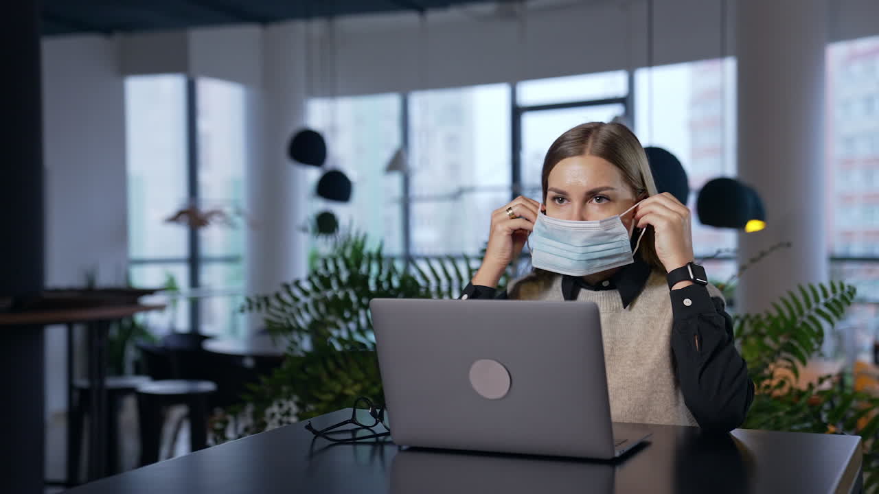 Girl in office puts a mask on and fits it to her face. Long-haired female employee wears mask and continues her work at the computer.