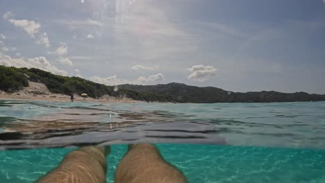 First-person-perspective-of-man-legs-and-feet-floating-on-beautiful-clear-and-transparent-sea-water-of-turquoise-lagoon-of-Saleccia-beach-in-Corsica-island,-France