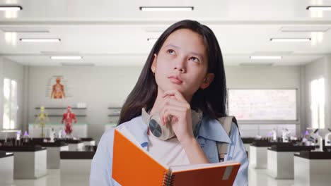 close up of asian teen girl student with a backpack reading book and thinking and looking around then raising her index finger while standing in science laboratory