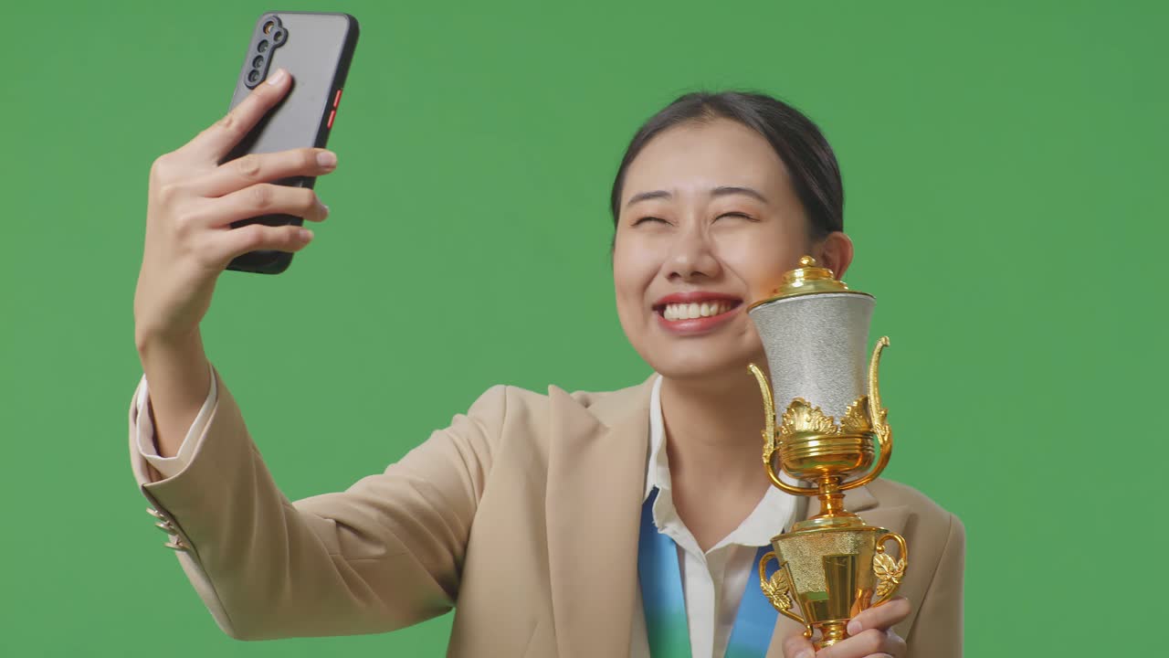 금메달을  슈트 입은 아시아 사업가 여성이 스마트폰으로 사진을 찍으며 스튜디오에서 초록색 화면 배경에서 첫 번째 우승자로 사진을 촬영합니다.