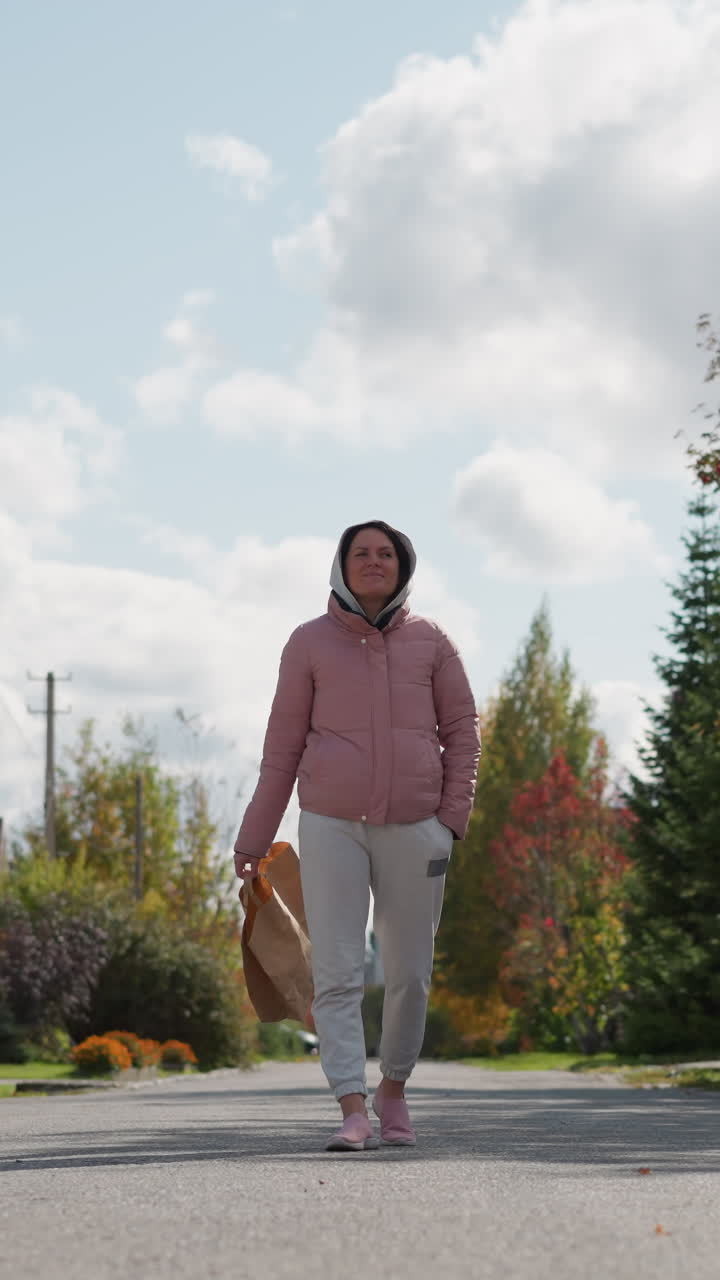 mujer de negocios en chaqueta rosada y pantalones de deporte grises se aleja con una bolsa de papel balanceándose por la calle residencial, rodeada de vibrante follaje de otoño y casas pintorescas bajo cielos brillantes