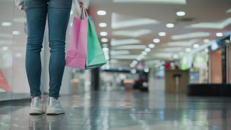 close-up of shopping bags held by a woman standing in a well-lit mall, the bags, one pink and the other green, sway gently as she walks on shiny flooring with blurred background