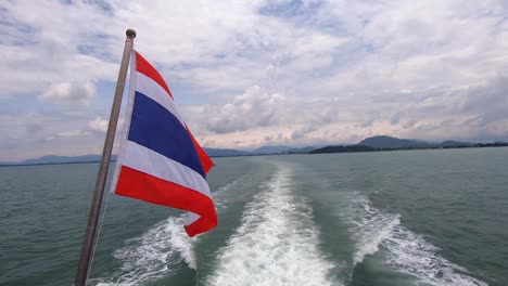thailand flag waving in wind on a boat ride to phi phi island with phuket in the background as boat wake pushes through ocean water in slow motion