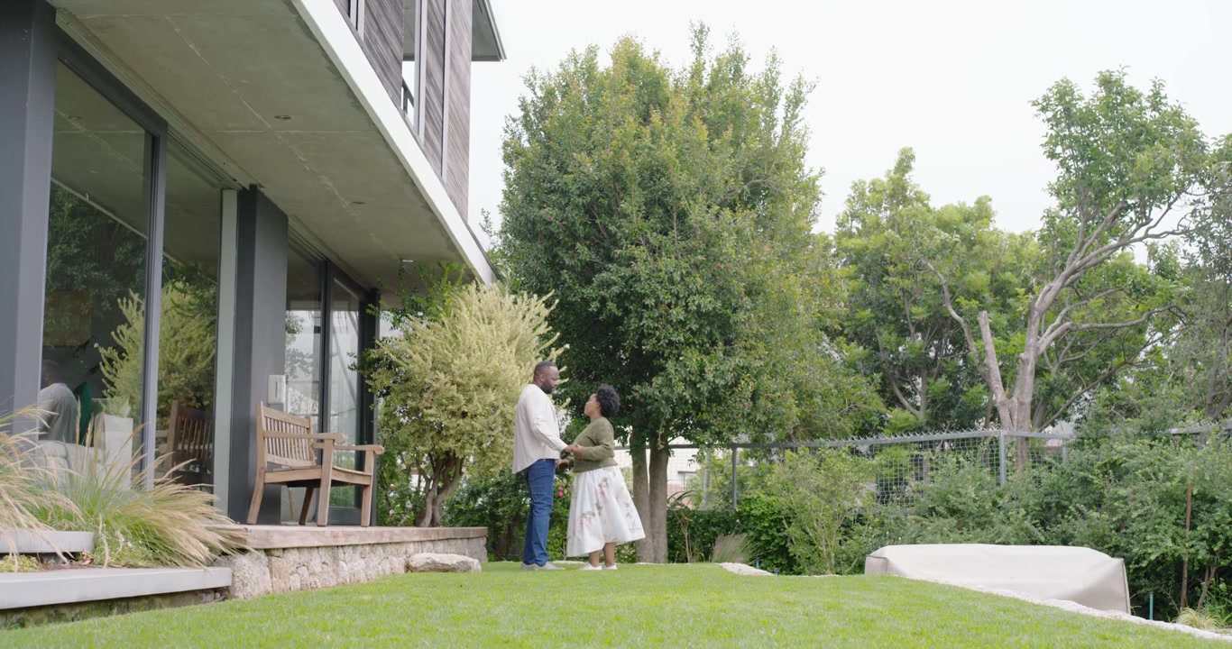 Coppia afroamericana che cammina tenendosi per mano, lasciando il patio di casa, fermandosi sotto un albero a conversare.
