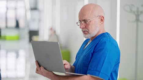 zoom-in-A-confident-elderly-man-with-glasses-a-doctor-in-a-blue-uniform-stands-in-the-middle-of-the-corridor-of-a-bright-clinic-and-types-on-his-gray-laptop