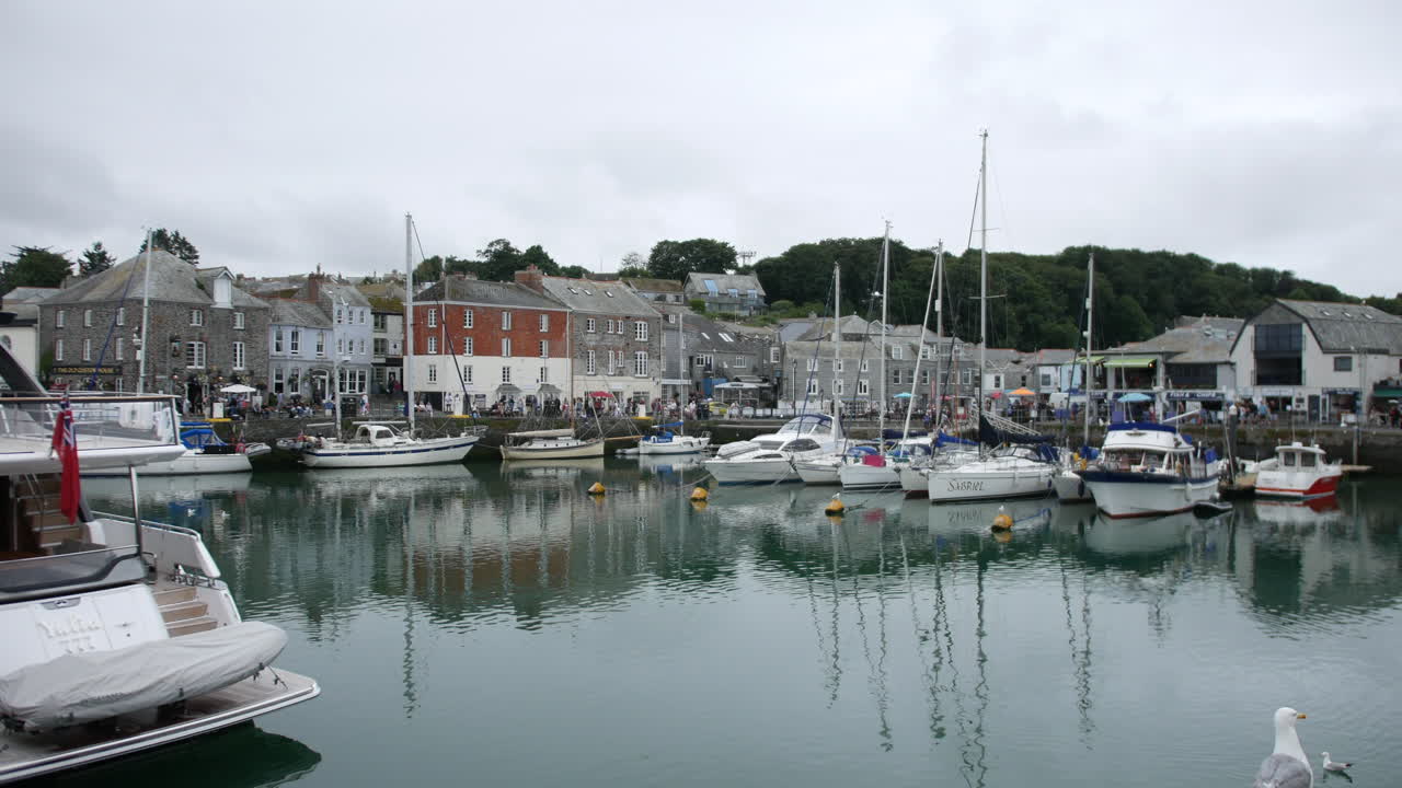 Sailboats and yachts anchored in a bustling harbor are surrounded by historic waterfront buildings and visitors exploring Padstow, Cornwall, England
