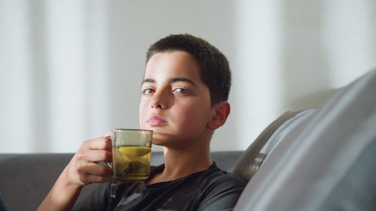 Boy sitting on a couch holding a tea cup close to his mouth, staring directly at the camera with a calm and focused expression, creating a serene and contemplative mood