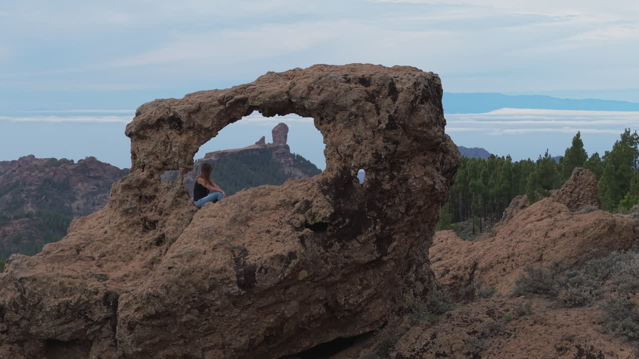 Aerial footage of a woman sitting in the Window of Roque Nublo (Gañifa), admiring the iconic Roque Nublo in the background. Gran Canaria, Canary Islands.