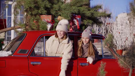 laughing teen girls looking out car window with decorative christmas tree on roof. happy young girls in red car with new year tree and gifts. expression positive emotion on christmas eve.