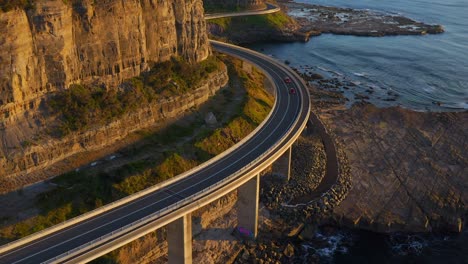 flying towards sea cliff bridge with cars driving on rocky coastline in northern illawarra region of new south wales, australia