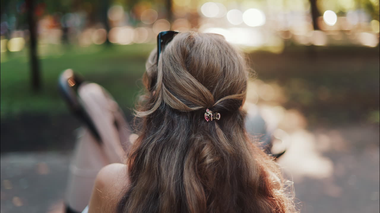 A mother with long wavy hair siting on a bench, breastfeeding her baby in a peaceful park, next to a modern baby stroller