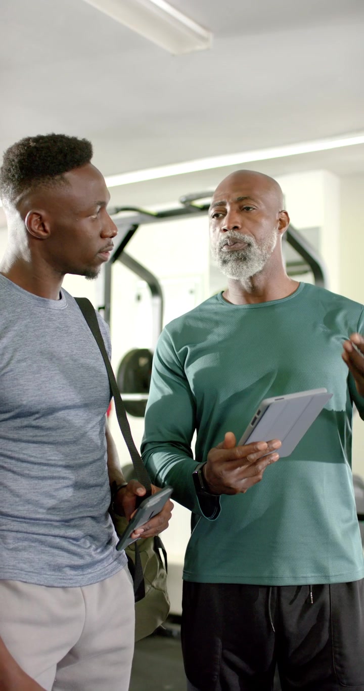 Verticaal video: Afro-Amerikaanse coach tikt op een tablet en wijst naar een rek, klant bestudeert telefoon in de sportschool