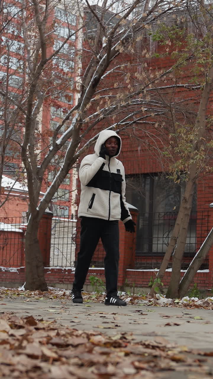 hombre africano teniendo una conversación telefónica mientras paseaba en un parque urbano durante el invierno, las hojas secas cubren el suelo con árboles y edificios borrosos en el fondo