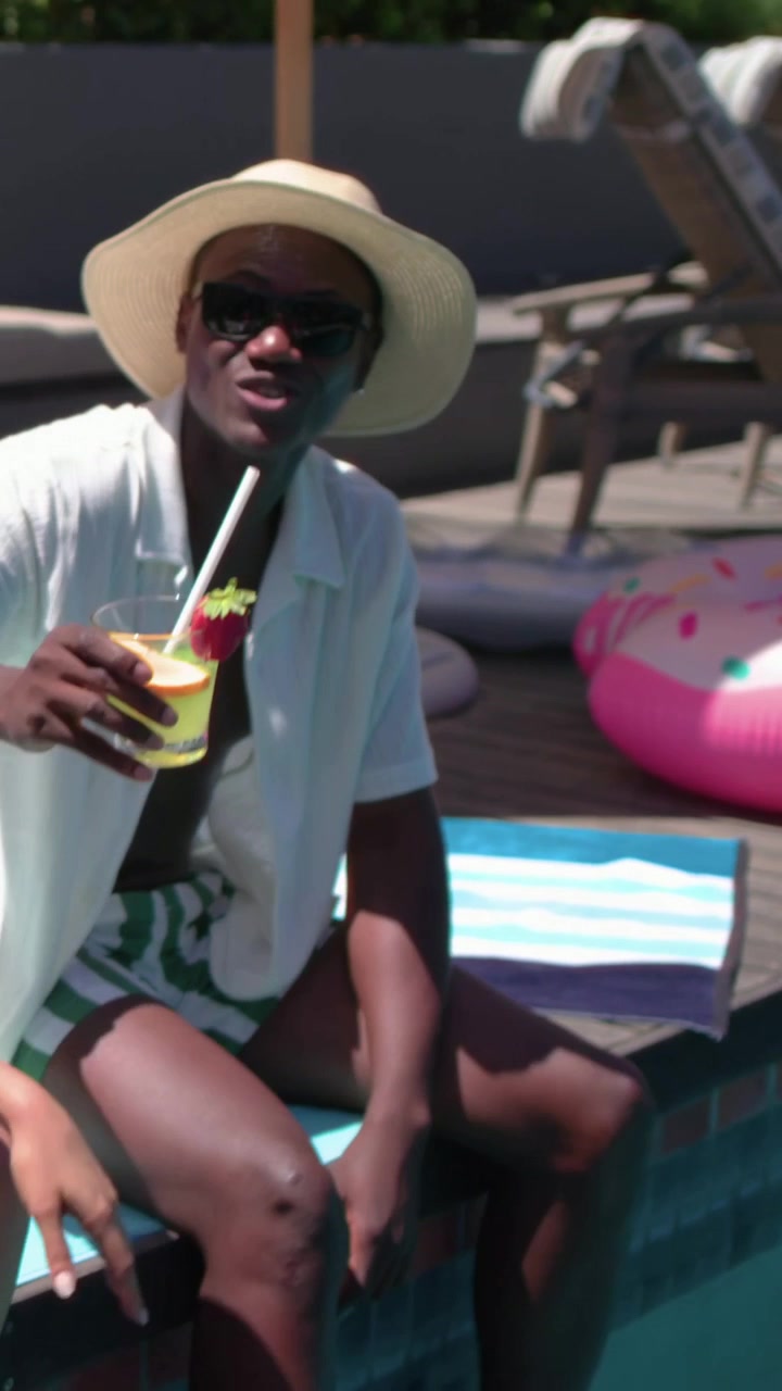 Video verticale: Uomo con cappello di paglia e occhiali da sole sorseggia un cocktail e alza il bicchiere a bordo piscina, al sole.