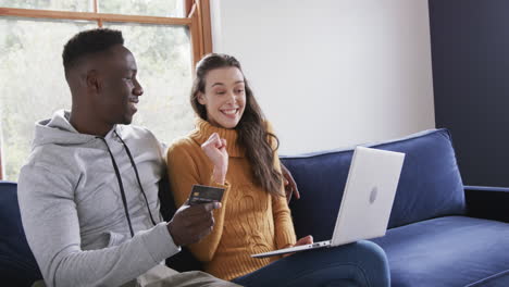 happy diverse couple sitting on sofa, using laptop and credit card in home,copy space