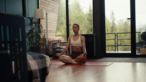 A-blonde-girl-in-a-white-top-with-tattoos-is-engaged-in-meditation-and-yoga-on-a-special-rug-in-a-modern-house-against-the-backdrop-of-a-green-forest-outside-the-window