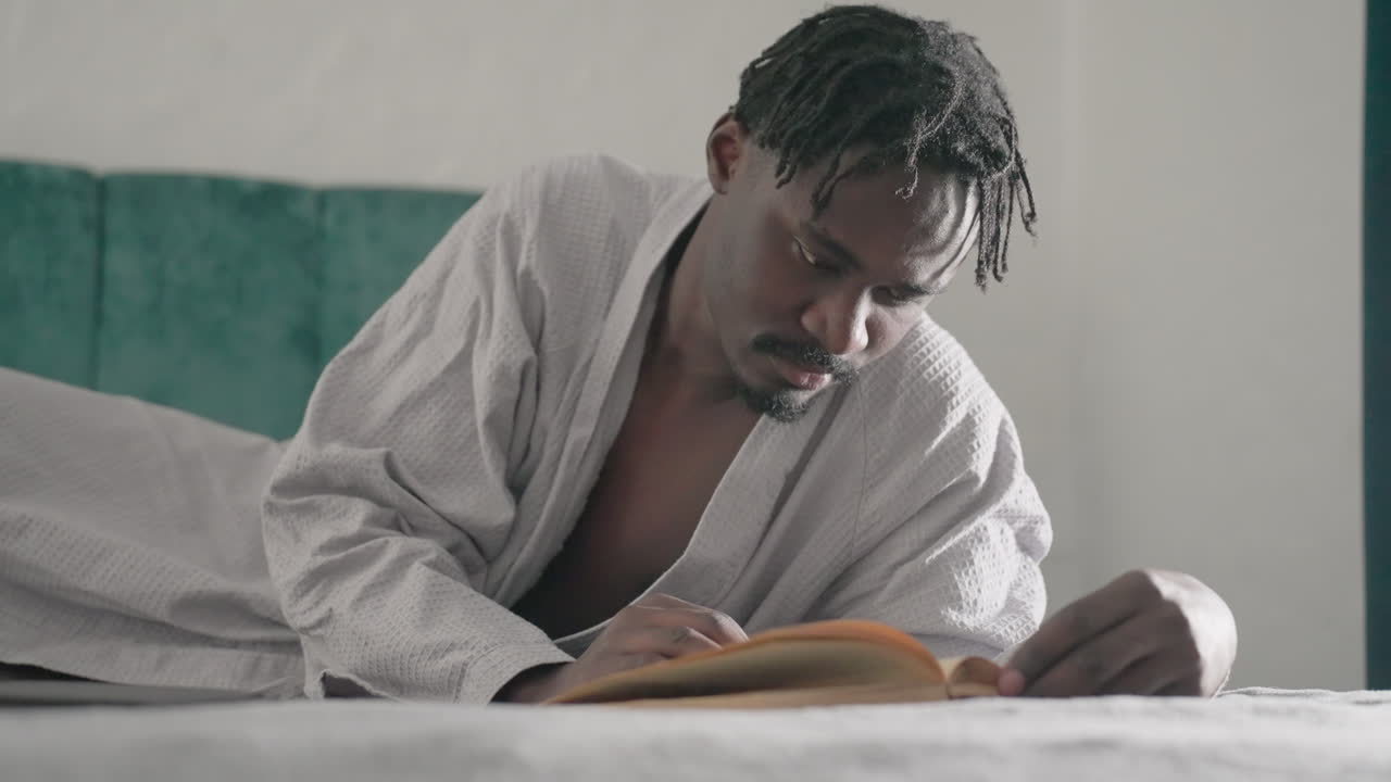 Man in white robe reading large book while lying on bed, focused on pages. Soft light filters through window, creating a peaceful atmosphere, as he turns the pages slowly, enjoying the content